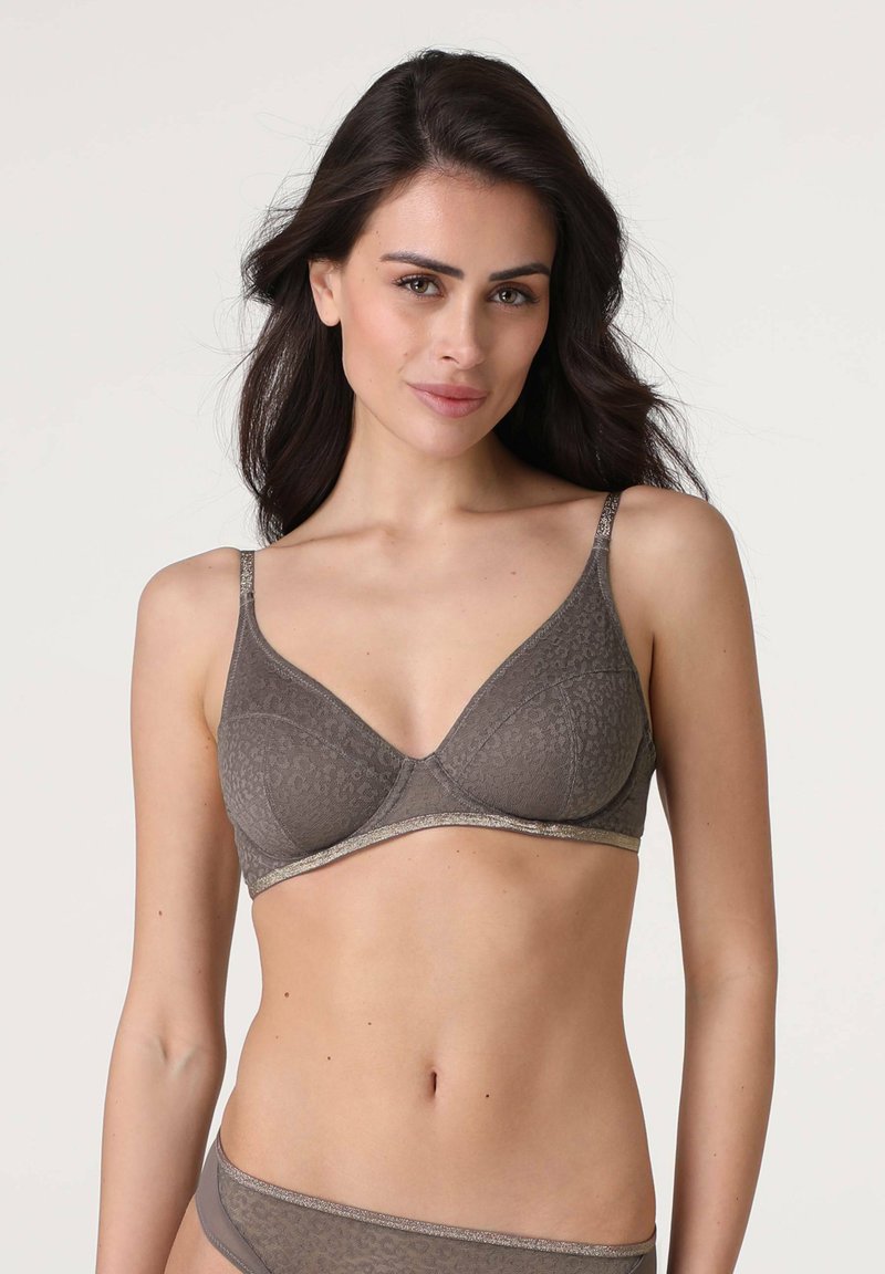 Donna che indossa un reggiseno a fantasia marrone e mutandine abbinate, in posa contro uno sfondo chiaro e semplice con capelli scuri e sciolti.