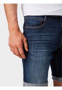 Tummansininen farkkushortsit käännetyllä helmalla, jossa on kevyt kuluminen ja erottuva ommel. Käsi lepää sivutaskulla.