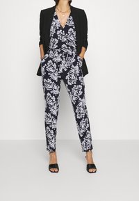 Woman wearing black blazer over navy floral jumpsuit with hands in pockets, black heeled sandals, and layered gold necklaces.