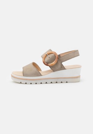 Beige suede wedge sandal with an open toe, curved ankle strap, and a large round buckle accent. White midsole with a textured outsole.