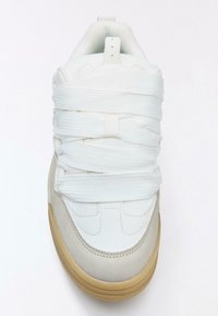 Fehér sportcipő texturált bőr testtel, rugalmas fehér fűzőkkel és bézs gumitalppal. Párnázott szegéllyel és varrt díszítésekkel rendelkezik.