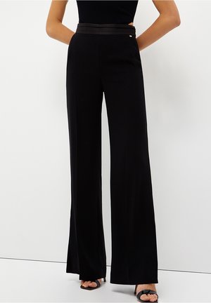 Pantalones negros de pierna ancha con una textura suave, con una cintura de doble capa y un sutil detalle metálico. Combinados con sandalias negras.
