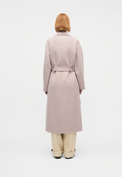 Άτομο με κοντά κόκκινα μαλλιά που φοράει μακρύ μπεζ παλτό με ζώνη και φαρδιά μπεζ παντελόνια, στέκεται με την πλάτη προς το φακό σε λευκό φόντο.