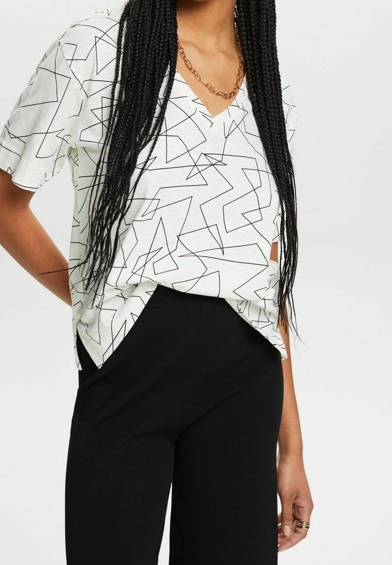 Femme portant un haut blanc à manches courtes avec des lignes géométriques noires, un pantalon noir, des cheveux tressés aux épaules et un collier chaîne doré.