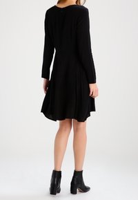 Svart långärmad klänning med en utsvängd kjol, tygstruktur synlig, med en nyckelhålsknäppning bak och bärs med svarta ankelstövlar.