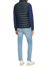 Gepolsterte marineblaue Weste mit gestepptem Muster, kombiniert mit einem blauen strukturierten Langarmshirt, hellblauen Jeans und weißen Turnschuhen.