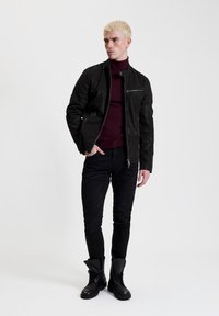 Giacca di pelle nera con zip frontale, dotata di una tasca sul petto sinistro. Indossata sopra un dolcevita bordeaux, abbinata a jeans slim-fit neri e stivali.