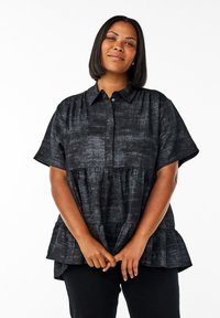 Woman with shoulder-length hair wearing a short-sleeve dark textured blouse with a collar, standing with hands clasped in front.