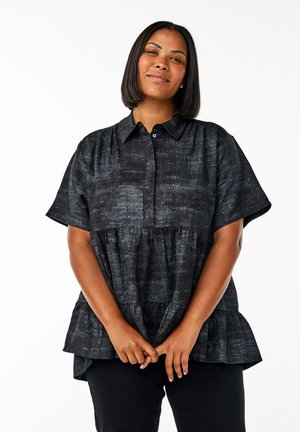 Woman with shoulder-length hair wearing a short-sleeve dark textured blouse with a collar, standing with hands clasped in front.