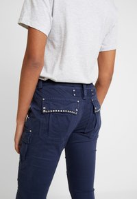 Granatowe spodnie cargo z teksturowanej tkaniny, z ćwiekowanymi akcentami na kieszeniach i dopasowanym krojem. Tylny widok ukazuje szczegóły kieszeni.