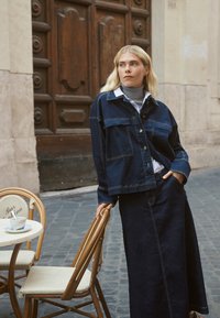 Jeansjacke mit Fronttaschen und Knopfverschluss, getragen über einem hellen Hemd mit Kragen, kombiniert mit einem langen, passenden Jeansrock. Dunkelblaue Farbe.