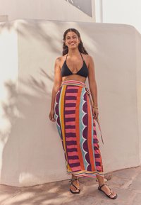 Black bikini top, long multicolored wrap skirt with bold stripes, scalloped patterns, and tied waist. Black flat sandals on stone surface.