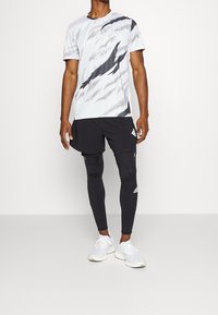 Męski strój sportowy składający się z T-shirta w biało-szary wzór oraz czarnych szortów, w zestawieniu z dopasowanymi czarnymi legginsami i białymi sneakersami.