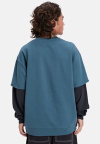 Sudadera de corte holgado y manga corta en color verde azulado, fabricada en una mezcla de algodón. Presenta un aspecto superpuesto con una camisa de manga larga negra debajo y puños acanalados.