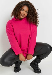 Sudadera de gran tamaño de color rosa intenso con un cuello alto, mangas largas y un corte relajado, combinada con leggings de cuero sintético negros y botas negras gruesas.