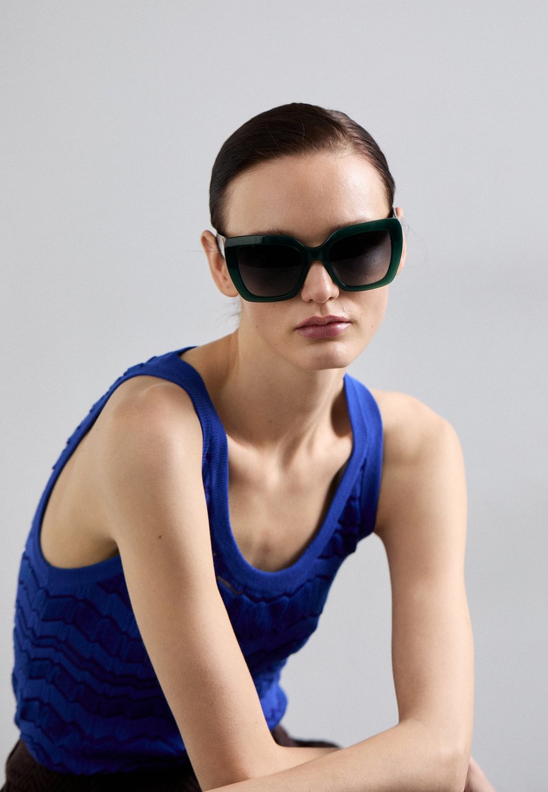 Sunglasses med en tjock grön ram och gradientlinser, kompletterade av en texturerad blå tanktopp med ett vågigt mönster.