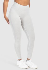 Donna che indossa leggings bianchi a vita alta e scarpe da ginnastica bianche, in piedi contro uno sfondo grigio chiaro uniforme.