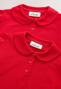 Dve crvene polo majice sa zaobljenim ovratnicima i tri dugmeta u obliku srca, a svaka ima Clarks oznaku marke unutar vrata.