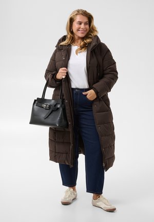 Femme portant un long manteau matelassé marron, une chemise blanche, un jean foncé et des baskets, tenant un sac à main noir avec une expression souriante.