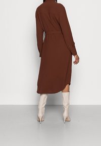 Robe longue marron avec une taille ceinturée, des poignets boutonnés et un pli au dos, associée à des bottes à talons crèmes hauteur genou. Texture lisse.