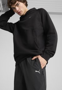 Melnā džemperis ar kapuci un kangaroo kabatu, ar smalku logo. Kombinēts ar tumši pelēkiem sašaurinātiem biksēm ar sānu rāvējslēdzēja kabatu.