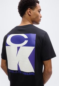 Mand iført en sort T-shirt med en stor blå og hvid CK Calvin Klein-logo trykt på ryggen, set fra siden.
