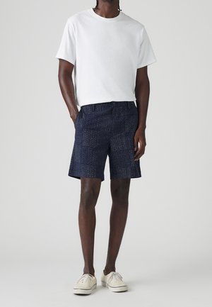 Homme portant un t-shirt blanc uni, un short à motifs bleu marine et des baskets crème, se tenant contre un fond gris clair.