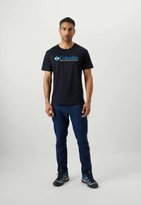 Fekete, rövid ujjú póló, rajta kék "Columbia Sportwear Company" logóval, kék cargo nadrággal és szürke túracipővel párosítva. Egyszerű dizájn.