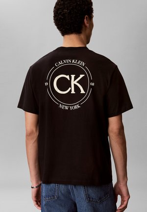 Mand iført sort Calvin Klein T-shirt med stort hvidt CK-logo, "Calvin Klein," "1968" og "New York" tekst på ryggen, kombineret med blå jeans.