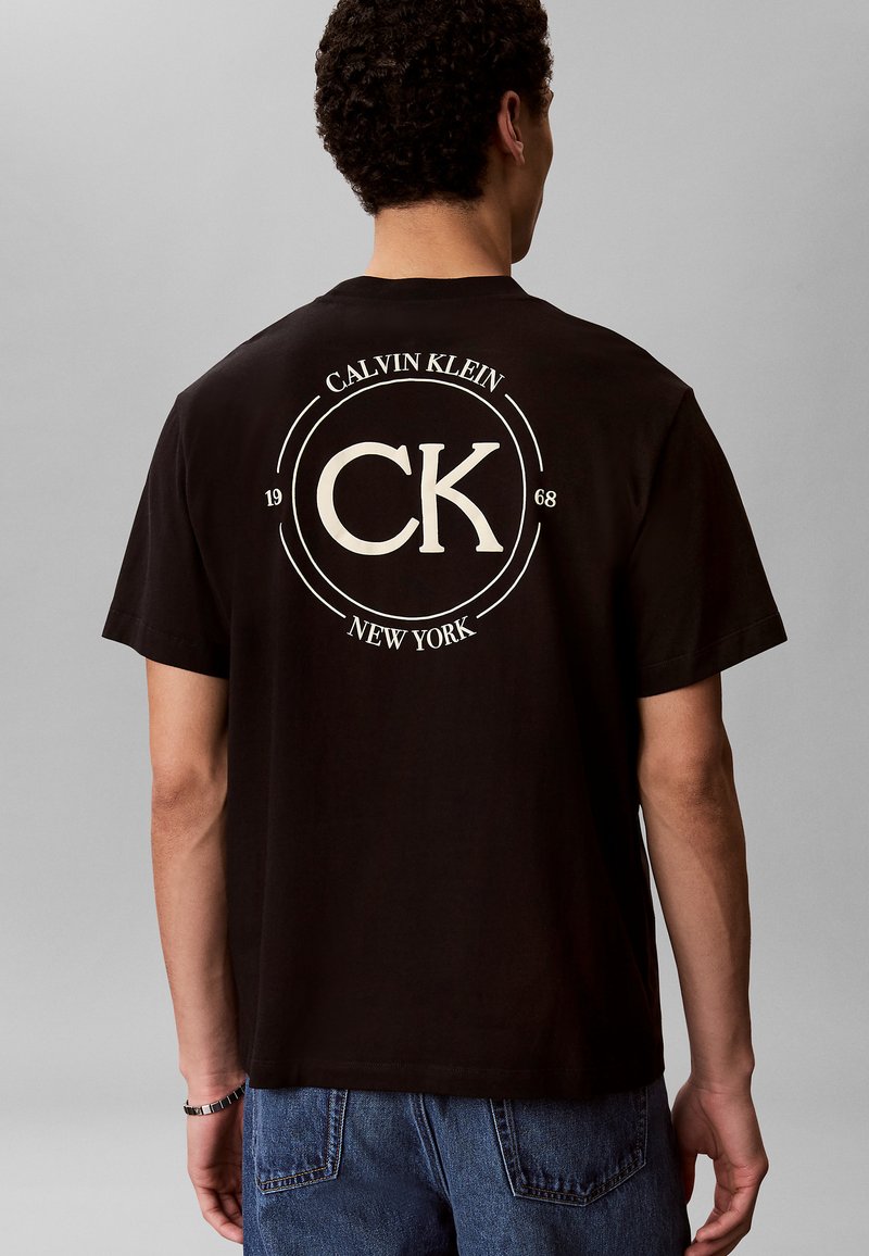 Homme portant un t-shirt noir Calvin Klein avec un grand logo CK blanc, les textes "Calvin Klein", "1968" et "New York" au dos, associé à un jean bleu.
