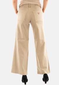 Pantalones de pierna ancha beige, hechos de algodón, que cuentan con dos bolsillos traseros y un pequeño logo. Textura suave con un ajuste relajado.