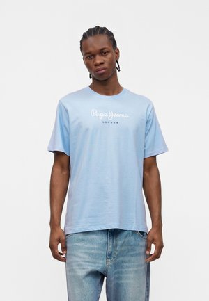 Młody mężczyzna z zaplecionymi włosami, ubrany w jasno niebieską koszulkę Pepe Jeans London i wyblakłe niebieskie dżinsy, stojący na jednolitym białym tle.