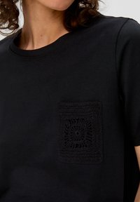 Zwarte t-shirt met een ronde halslijn, korte mouwen en een zwarte gehaakte zak met textuur en een bloemenpatroon.