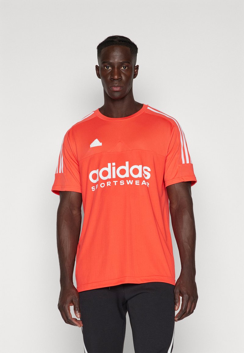 Camiseta deportiva roja de Adidas con logo blanco y franjas en las mangas. Presenta un cuello redondo y un diseño de tela texturizada. Pantalones negros puestos.