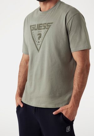 Mężczyzna w oliwkowej koszulce Guess z logo i czarnych spodniach z sznurkiem, ręce w kieszeniach, na tle jasnego, jednolitego tła.