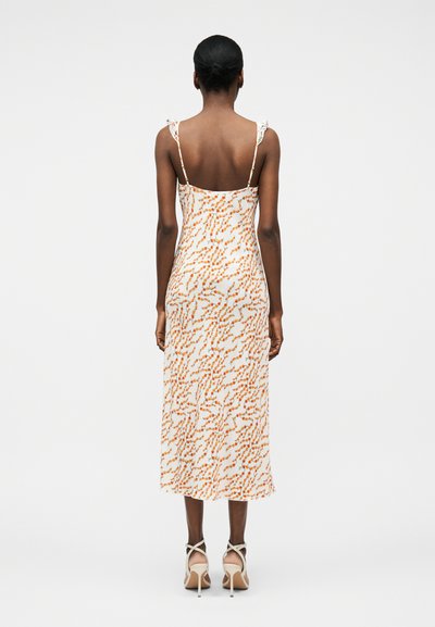 Γυναίκα που φοράει λευκό midi φόρεμα με πορτοκαλί λουλουδάτο μοτίβο, λεπτές τιράντες και ανοιχτή πλάτη, συνδυασμένο με λευκά σανδάλια με ψηλό τακούνι, στέκεται γυρισμένη προς τα πίσω.