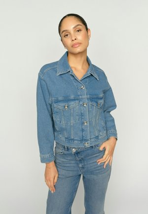Frau mit zurückgebundenem Haar, die eine blaue Jeansjacke und Jeans trägt, steht mit einer Hand in der Tasche vor einem einfarbigen hellen Hintergrund.