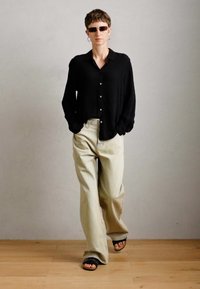 Personne aux cheveux courts portant des lunettes de soleil noires, un haut à manches longues noir, un pantalon large beige et des sandales noires, marchant les mains dans les poches.