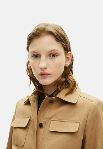 Veste beige à boutons avec col, deux poches avant et tissu texturé, présentant un design simple et une couleur neutre.