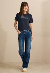 Camiseta navy con estampado "PIU AMORE", combinada con unos vaqueros de corte holgado en azul y zapatillas blancas con suelas beige sobre un fondo neutro.