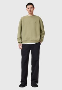 Sudadera verde oliva con un corte oversized, puños acanalados y un cuello redondo. Lleva puesta sobre una camiseta blanca, combinada con pantalones negros holgados y zapatos negros.