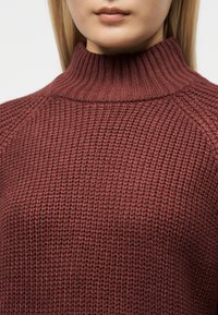 Gros plan sur une personne portant un pull bordeaux texturé, côtelé à col montant, mettant en évidence le motif tricoté et les détails du col.