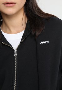 Sudadera negra con cremallera fabricada en una mezcla de algodón; presenta una cremallera plateada y el logotipo blanco de "Levi's" en el lado izquierdo del pecho. Capa interior blanca lisa visible.