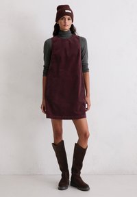 Burgundowa sukienka z korduroy z wysokim dekoltem, noszona na szarym golfie, w połączeniu z brązowymi kozakami do kolan i brązową czapką beanie.