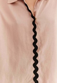 Svetloružová košeľa s textúrovaným čiernym vlnkovaným lemom pozdĺž predného zapínania so zlatými gombíkmi. Hladká látka, s golierom.
