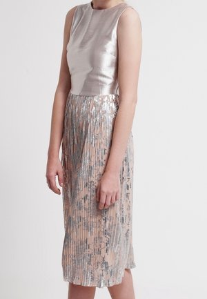 Vrouw draagt een mouwloos zilveren topje en een geplooide, metallic roze rok met zilveren bloemenpatronen, staand tegen een effen lichte achtergrond.
