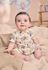 Gaiši bēšs jaundzimušo kombinezons ar krāsainām dzīvnieku un ēku ilustrācijām. Tam ir apkakle, īsas piedurknes un koka pogu aizdare.