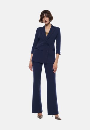 Blazer navy doppiopetto con maniche 3/4, abbinato a pantaloni a palazzo coordinati. Tessuto morbido, vestibilità sartoriale e décolleté a punta.