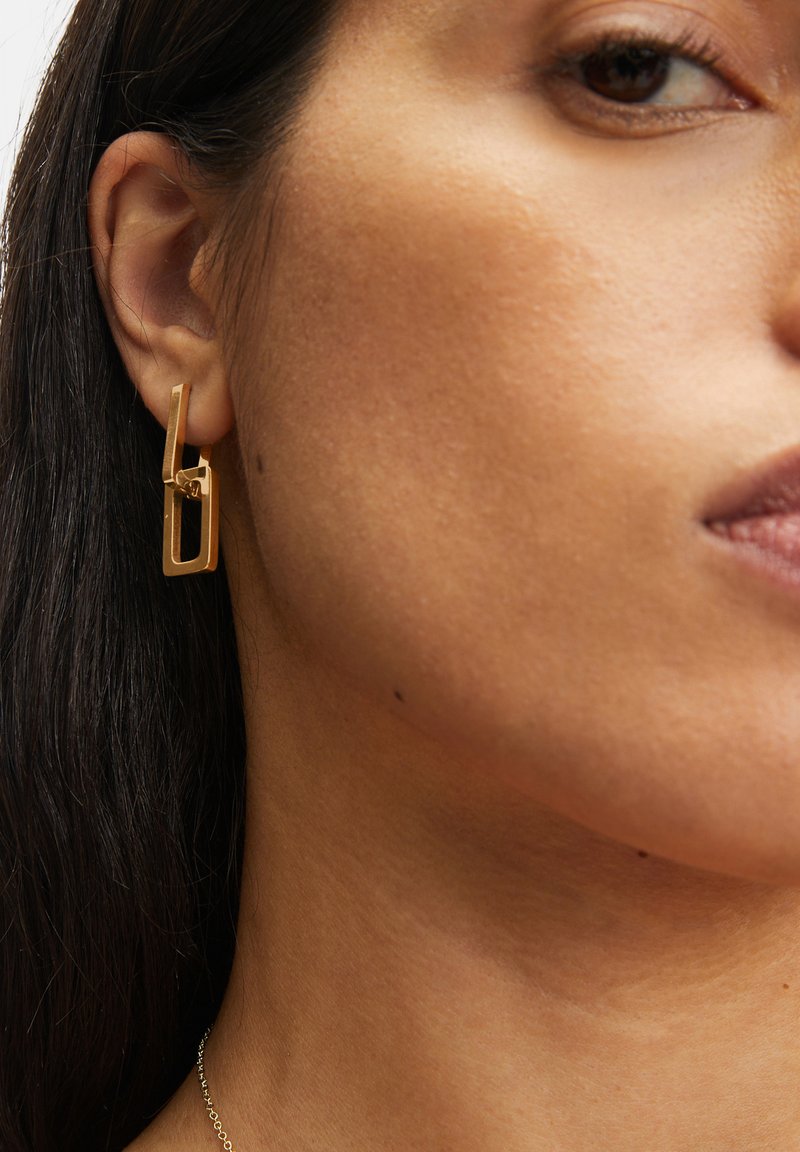Guld-farvede dangle øreringe har et geometrisk design med sammenflettede rektangulære former og en poleret overflade.