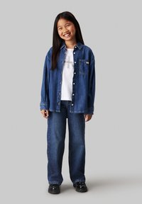 Denim shirt with button closure, long sleeves. Worn over a white graphic T-shirt and paired with blue wide-leg jeans. Black chunky shoes.